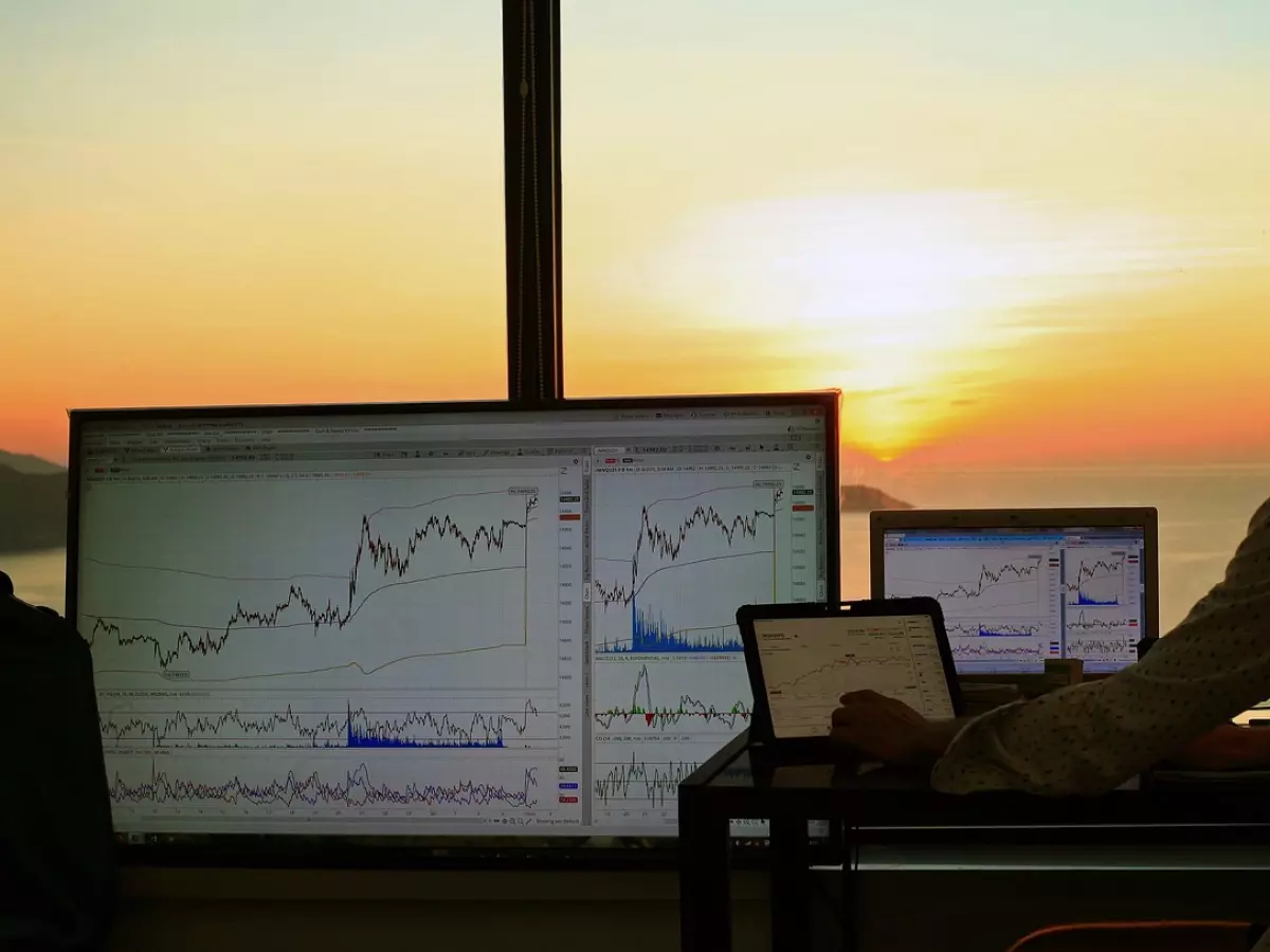 A man sits at a desk facing multiple monitors displaying financial charts. The sun sets in the background, casting a warm glow. 